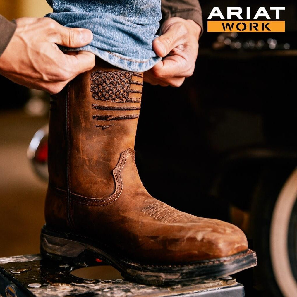 A person wearing a brown leather work boot with an American flag graphic near the top, standing on a small stool in a workshop setting. The person is pulling up blue jeans, and the Ariat Work logo is visible in the top right corner.